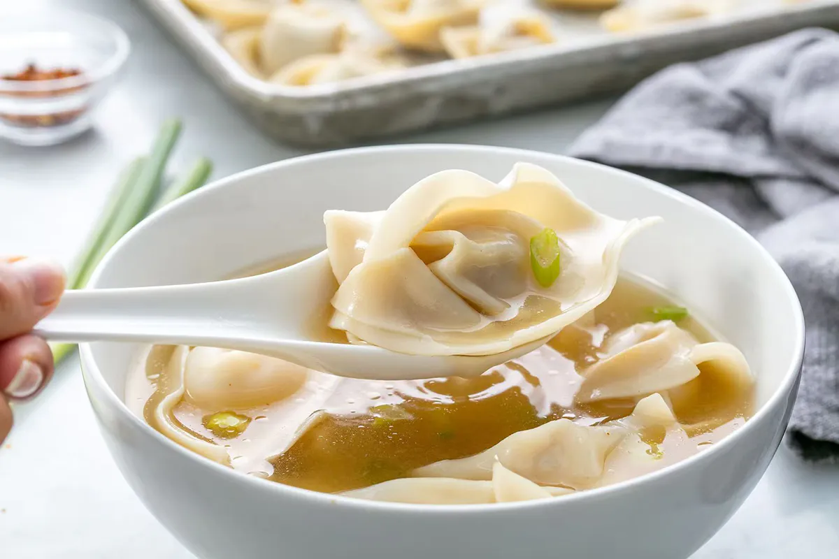 Wonton soup in white bowl, a wonton lifted on a spoon with green onion garnish at One More Bite Dumpling, a Chinese Restaurant in Palm Desert